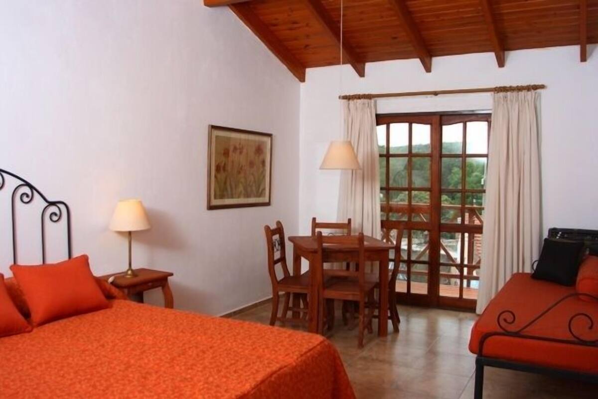 A room with a bed, a table with chairs, a window with curtains, and a painting on the wall.