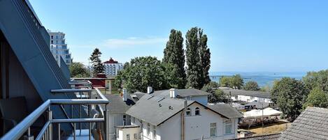 A residential area with a view of the sea, a balcony with metal railings, and a variety of buildings including a white house and a multi-story building.