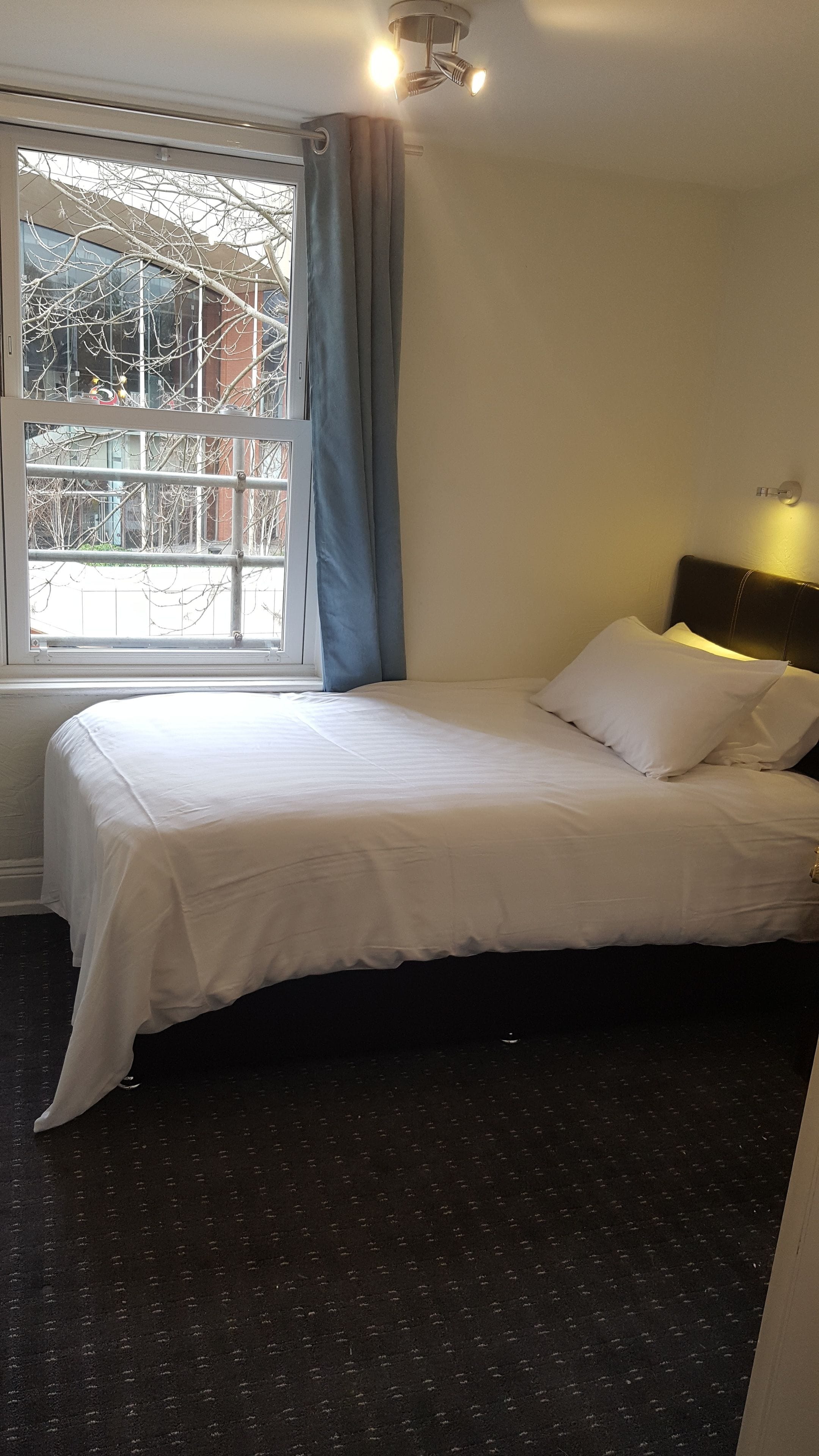 A hotel room with a bed, a window with curtains, a headboard, and a wall-mounted light fixture.