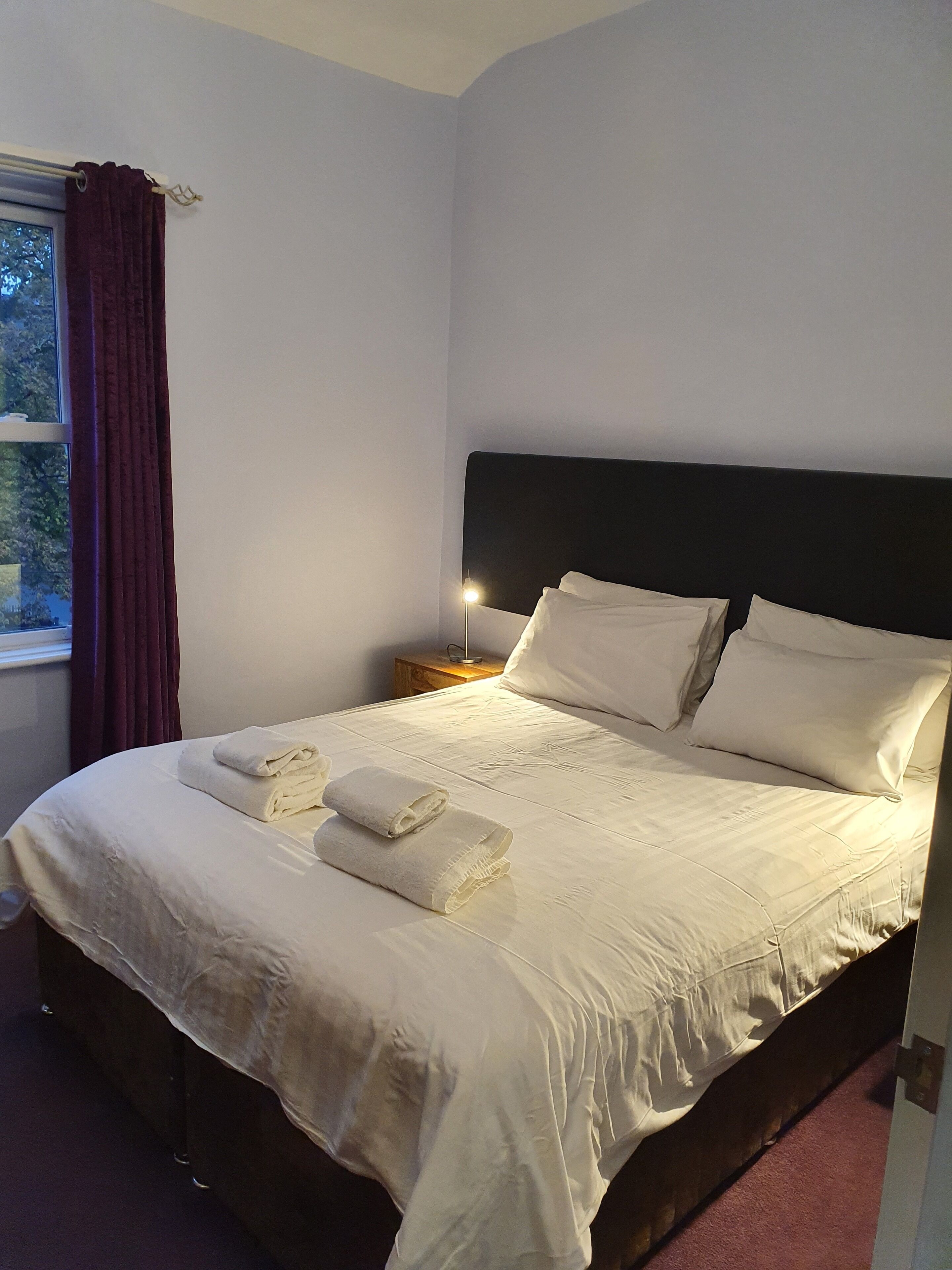 Una cama bien hecha con ropa de cama y toallas blancas, un cabecero oscuro y una mesita de noche con una lámpara.