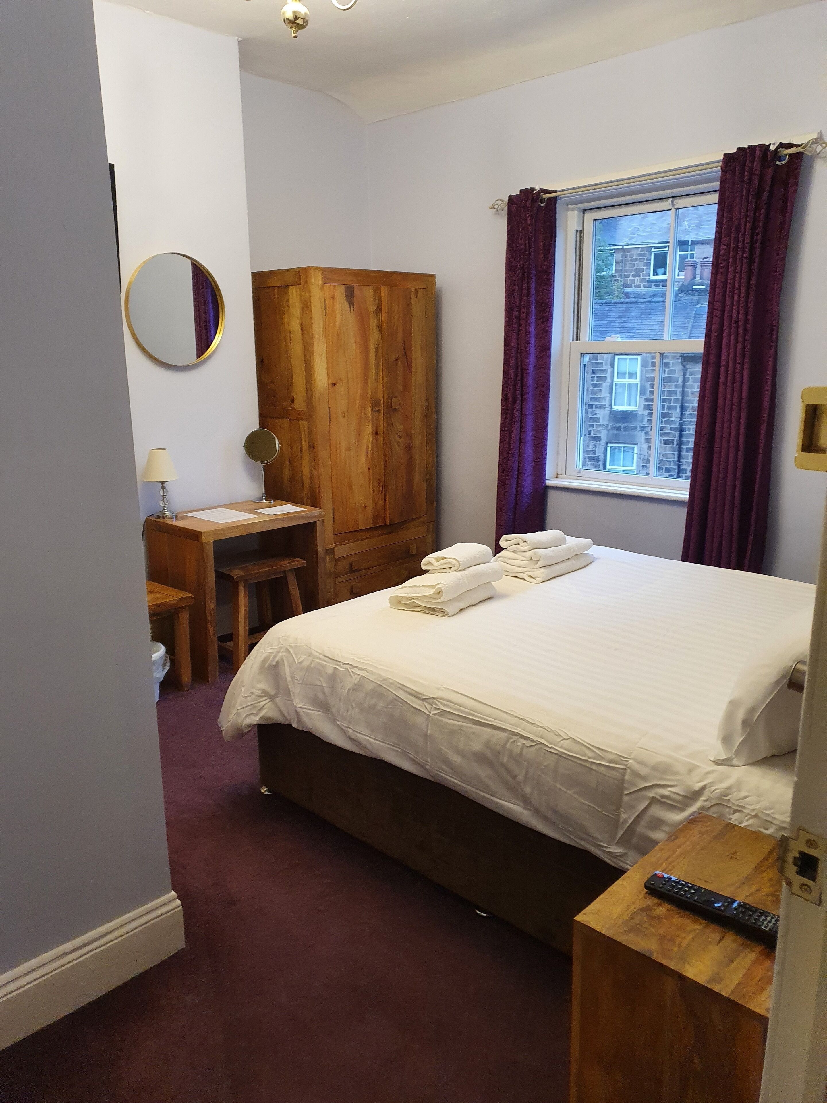 A bedroom with a bed, a wooden wardrobe, a desk with a lamp, a mirror, and a window with curtains.