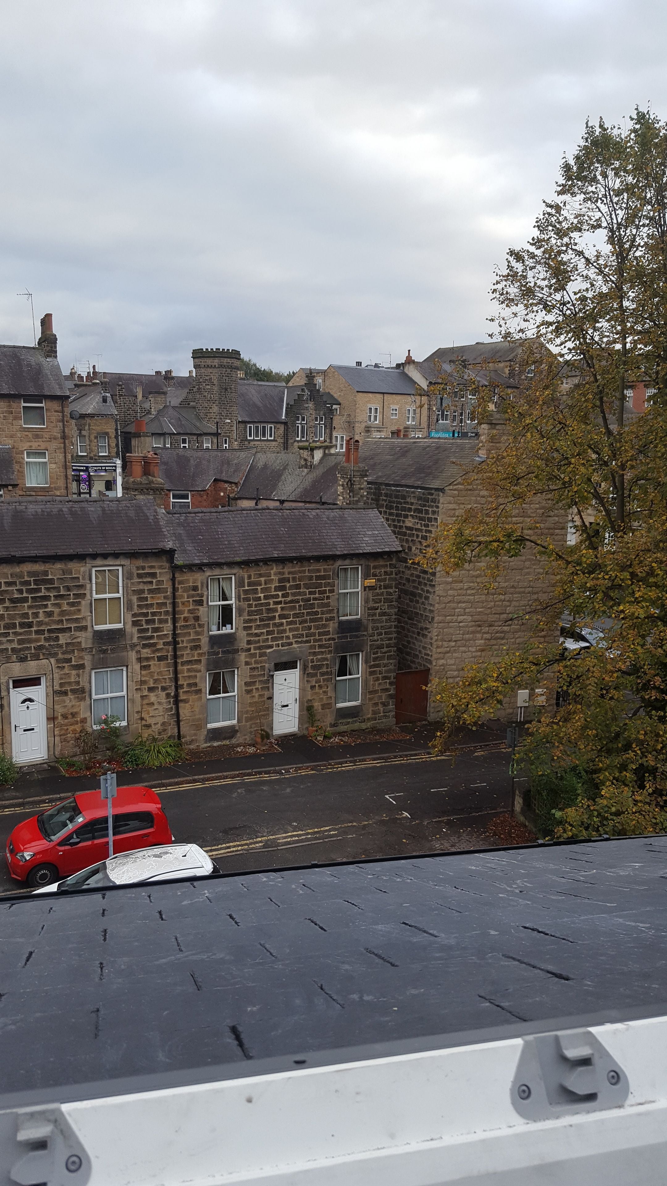 A view of a residential area with stone buildings, a red car parked on the street, and a tree in the foreground.