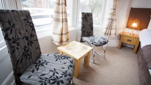 Habitación de hotel con una mesa de madera, dos sillas estampadas, una lámpara de noche y vistas al exterior a través de amplios ventanales.