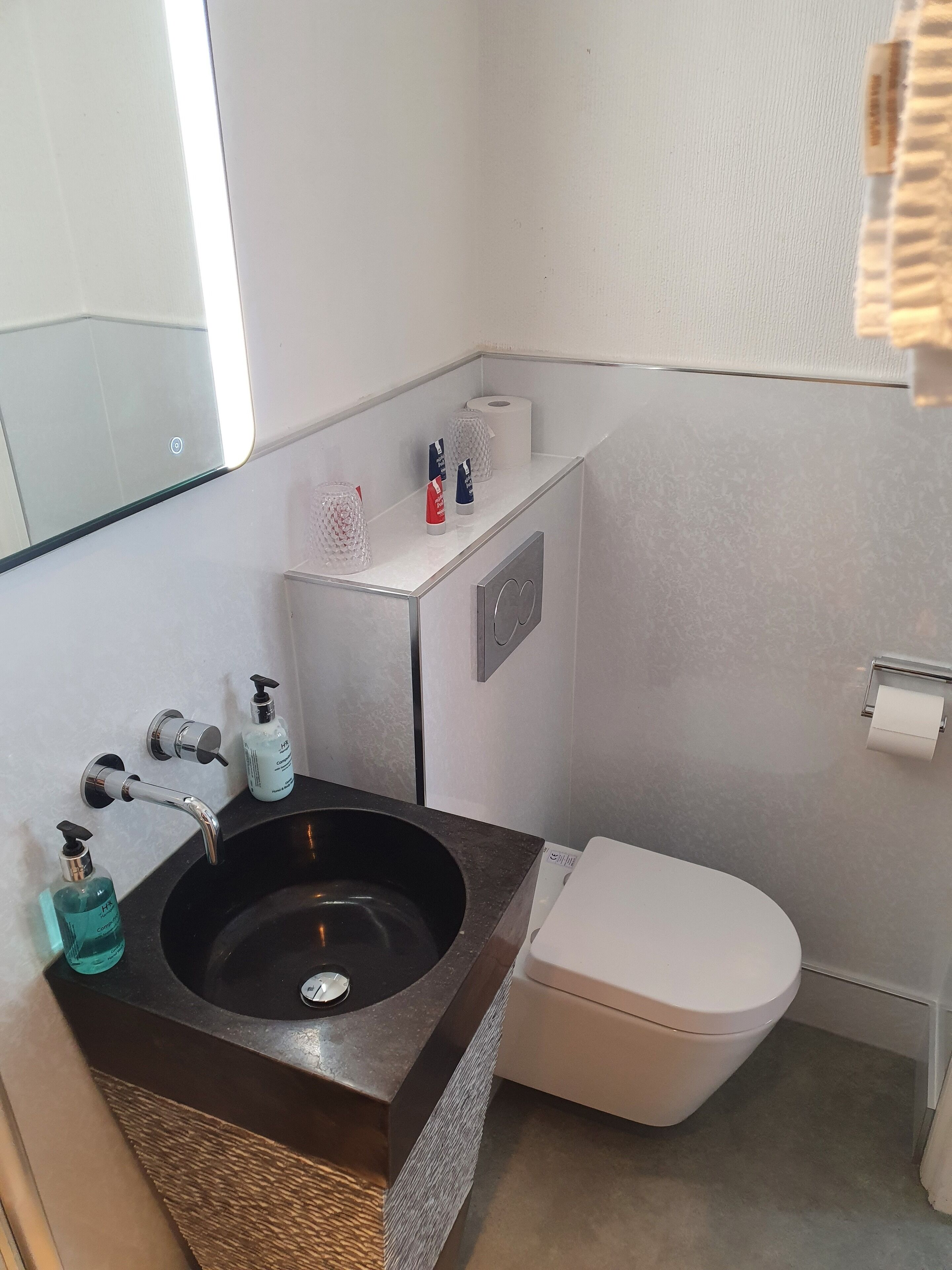 A bathroom with a black sink, white toilet, and a mirror above the sink. There are toiletries on the sink and a towel rack on the wall.