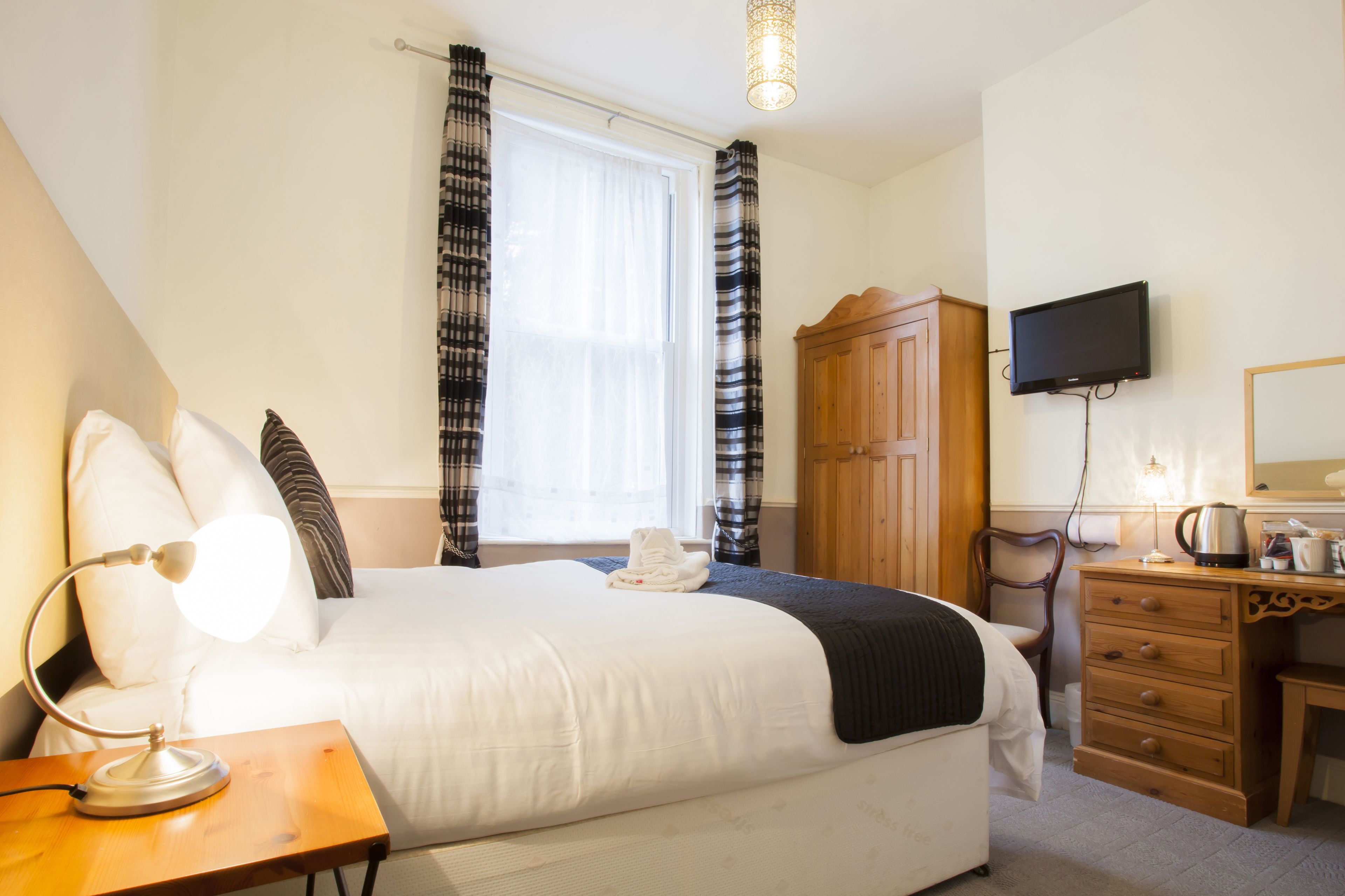 A hotel room with a large bed, a wooden dresser, a television mounted on the wall, and a window with curtains.