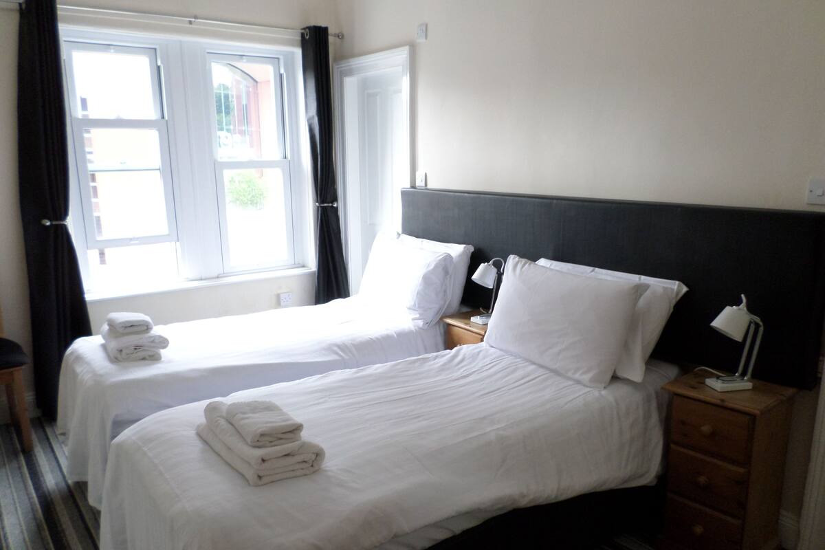 A hotel room with two single beds, a nightstand with a lamp, a wooden dresser, and a window with curtains.