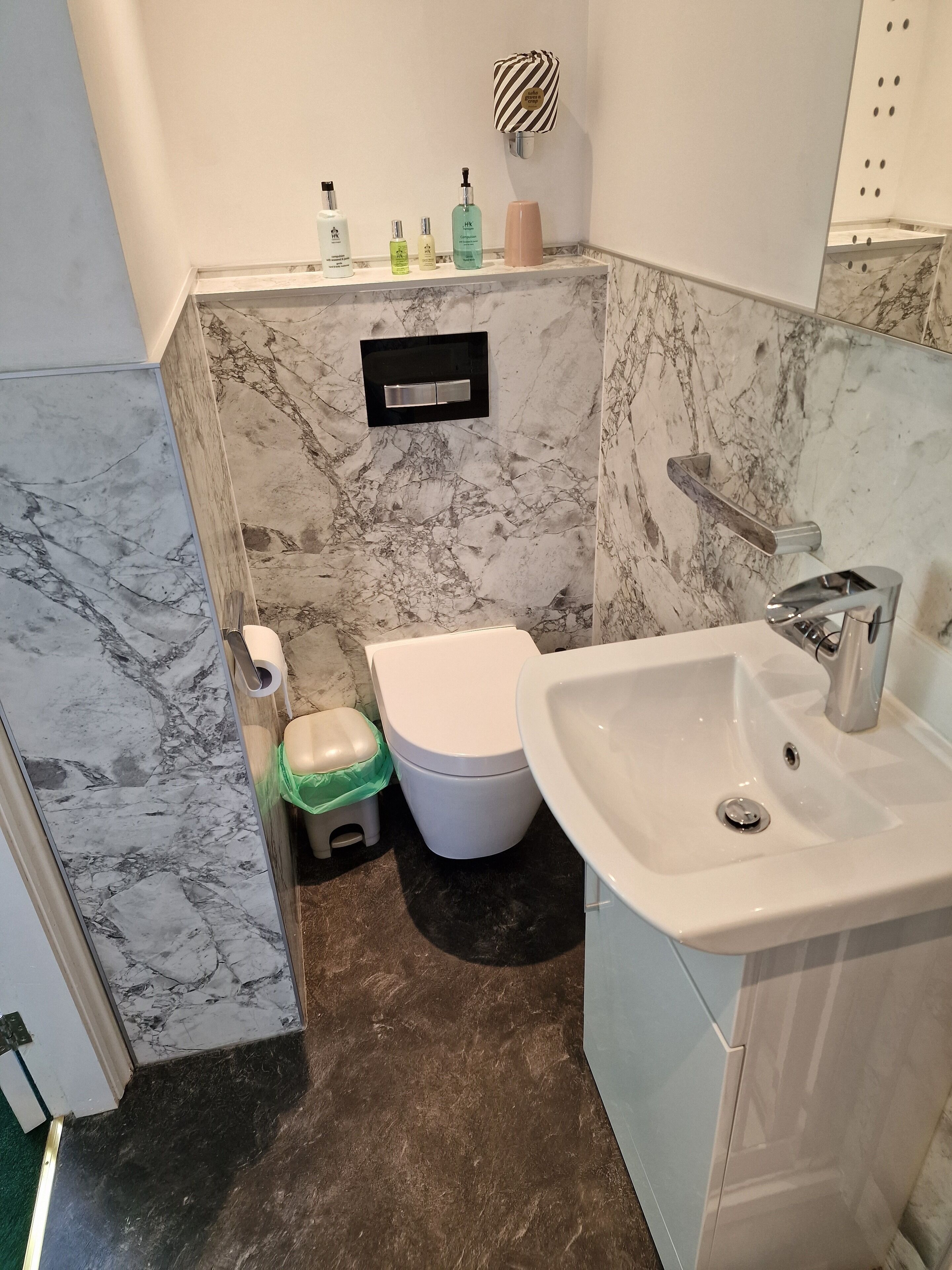 Un baño con suelo de mármol, inodoro blanco, lavabo y un secamanos en la pared.