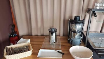 Preparación para el desayuno con cafetera, hervidor, cesta de utensilios, plato y un bol sobre una mesa de madera.