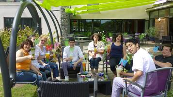 A group of people sitting at an outdoor table with drinks, surrounded by wicker chairs and a green canopy.