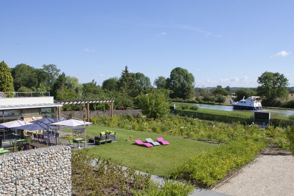 A riverside garden with outdoor seating, a boat on the water, and a modern building with a glass facade.