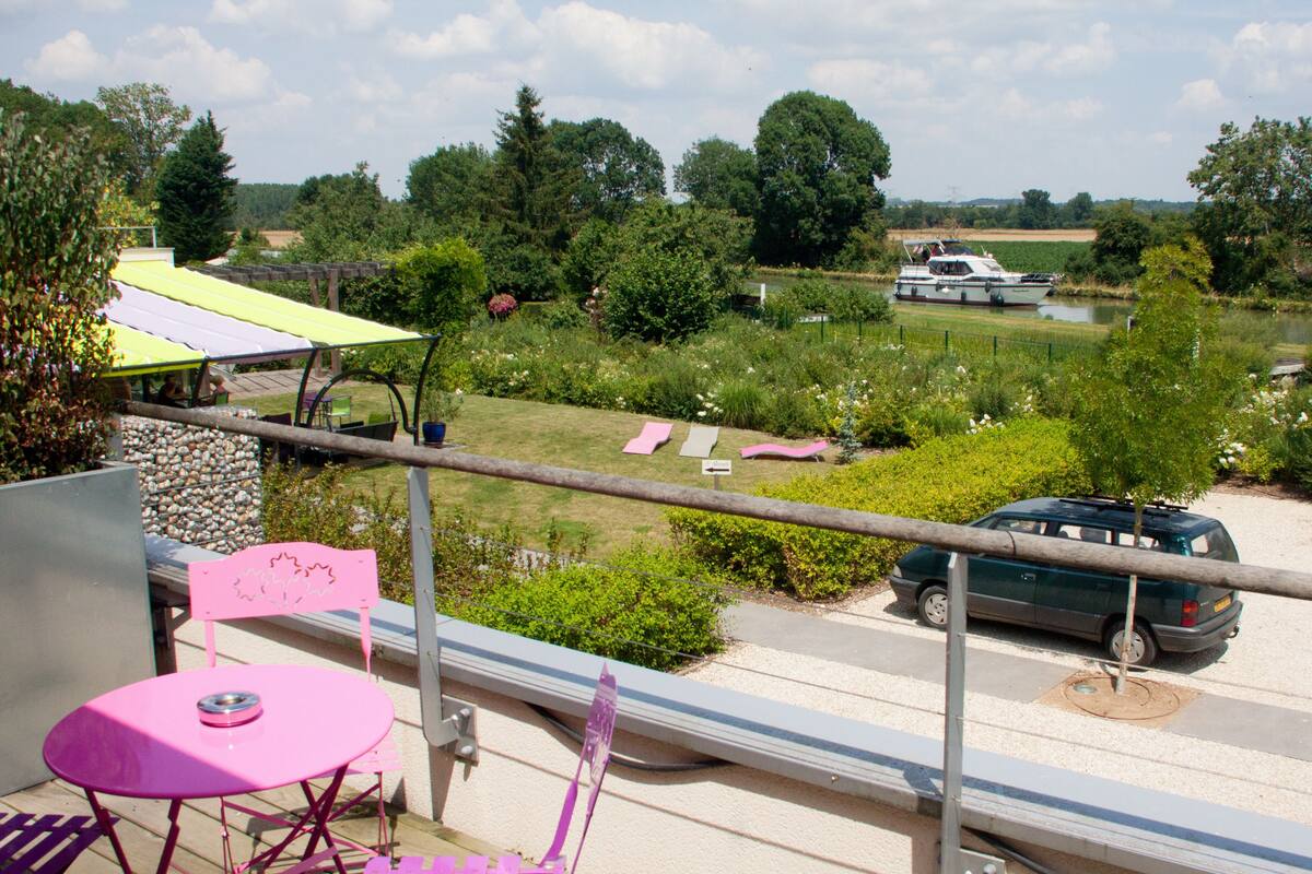 A balcony with a pink table and chair, overlooking a garden with a boat on a lake, and a parked car.