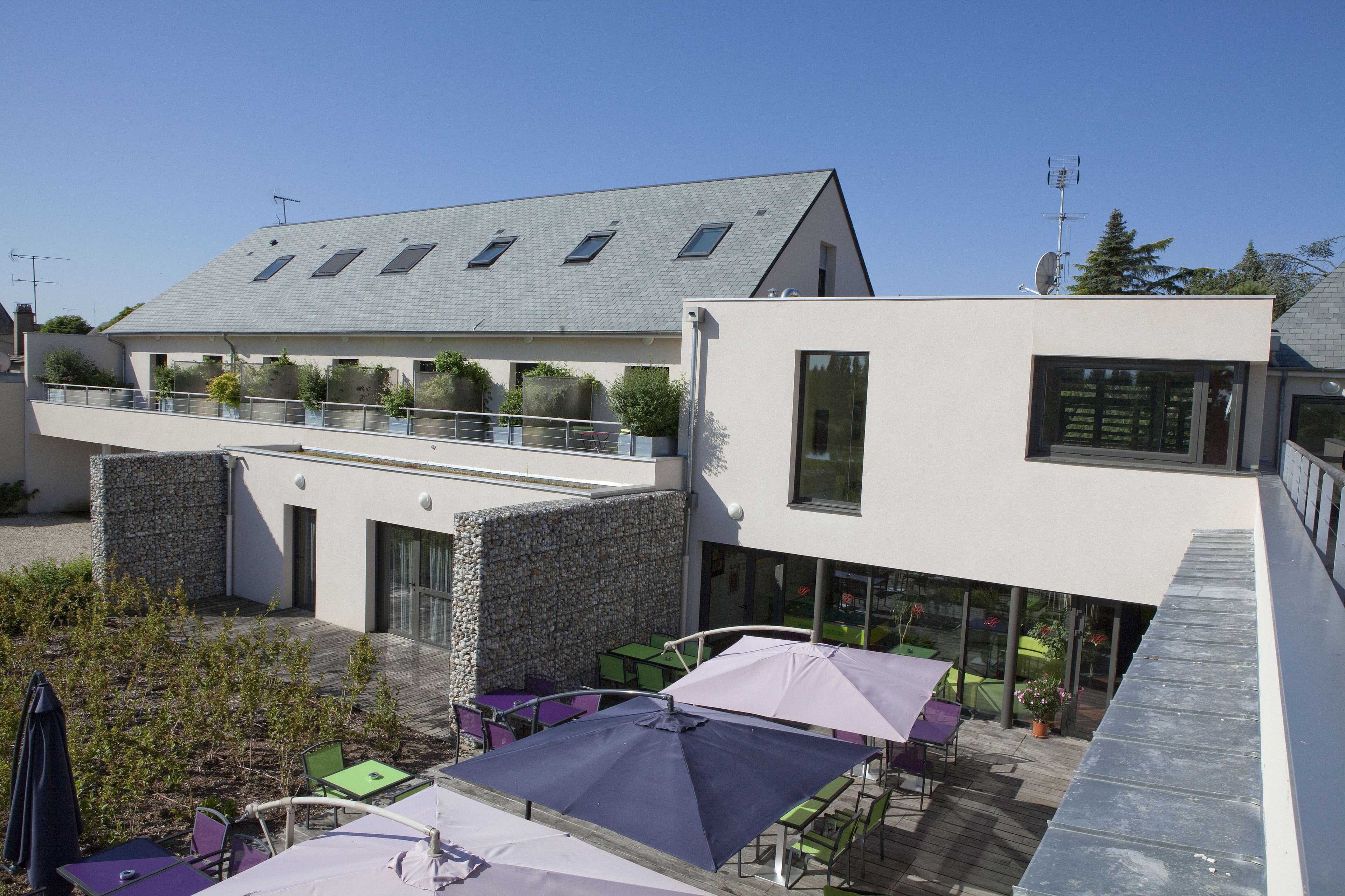 Ein modernes Gebäude mit flachem Dach, einem Balkon mit Glasgeländern und Sitzgelegenheiten im Freien mit Sonnenschirmen.