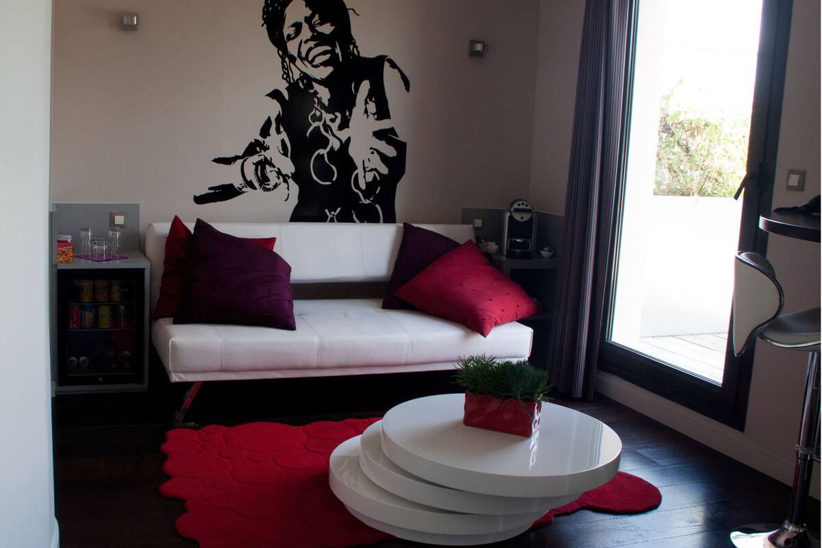 A modern living room with a white sofa, red cushions, a coffee machine, and a large window with curtains.