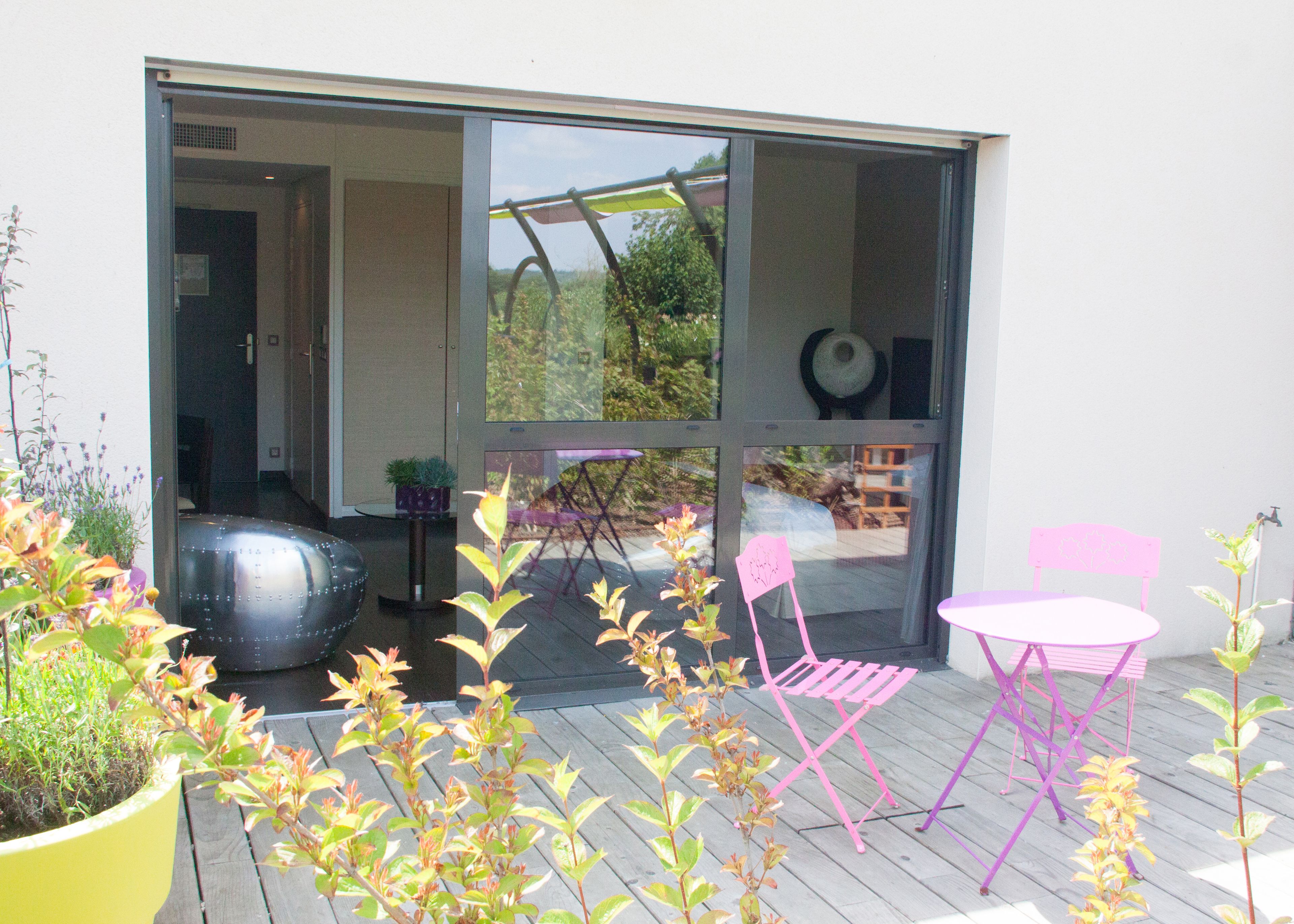An outdoor patio area with a glass door leading to a garden, a round table, two chairs, and potted plants.