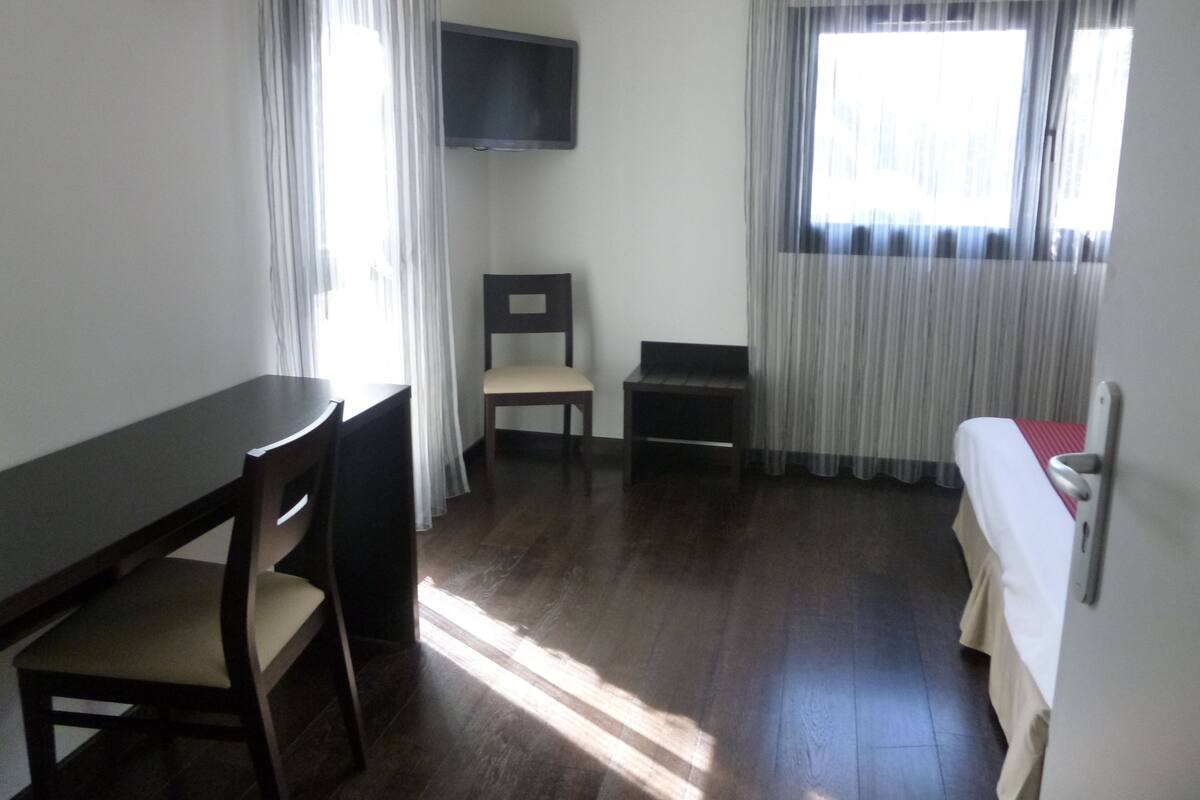 A room with a desk, two chairs, a television mounted on the wall, and a bed with a red blanket.