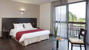 A hotel room with a large bed, a bedside table with a bottle and glasses, a glass table with fruit, a chair, and a view of the outdoors through a large window.