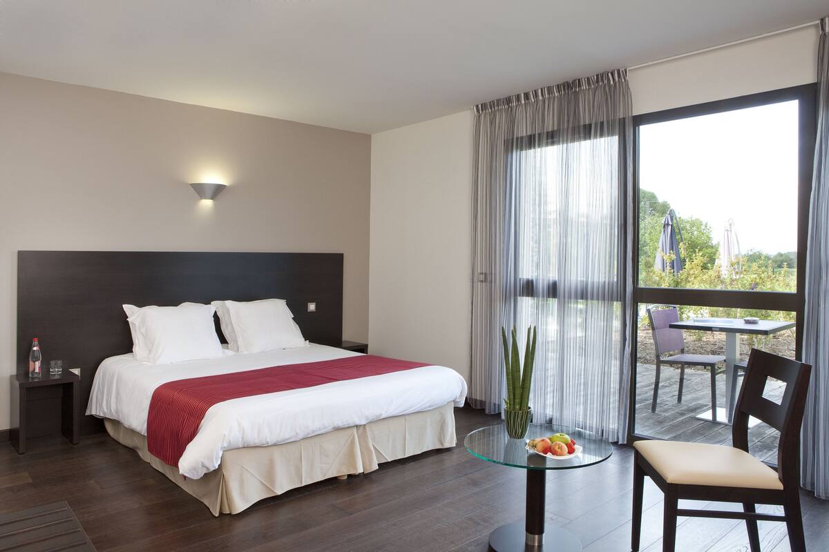 A hotel room with a large bed, a bedside table with a bottle and glasses, a glass table with fruit, a chair, and a view of the outdoors through a large window.