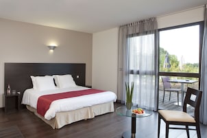 A hotel room with a large bed, a bedside table with a bottle and glasses, a glass table with fruit, a chair, and a view of the outdoors through a large window.