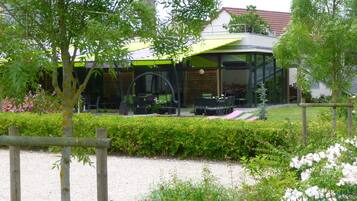 Un bâtiment moderne doté d’une façade en verre incurvée, d’un espace extérieur aménagé pour s’asseoir et d’un jardin bien entretenu avec des haies et des fleurs.