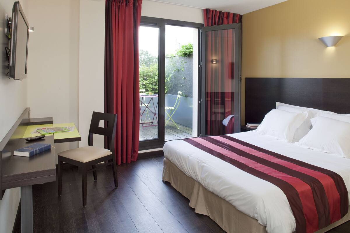 A hotel room with a bed, a desk, a chair, a TV, and a sliding glass door to the outside.