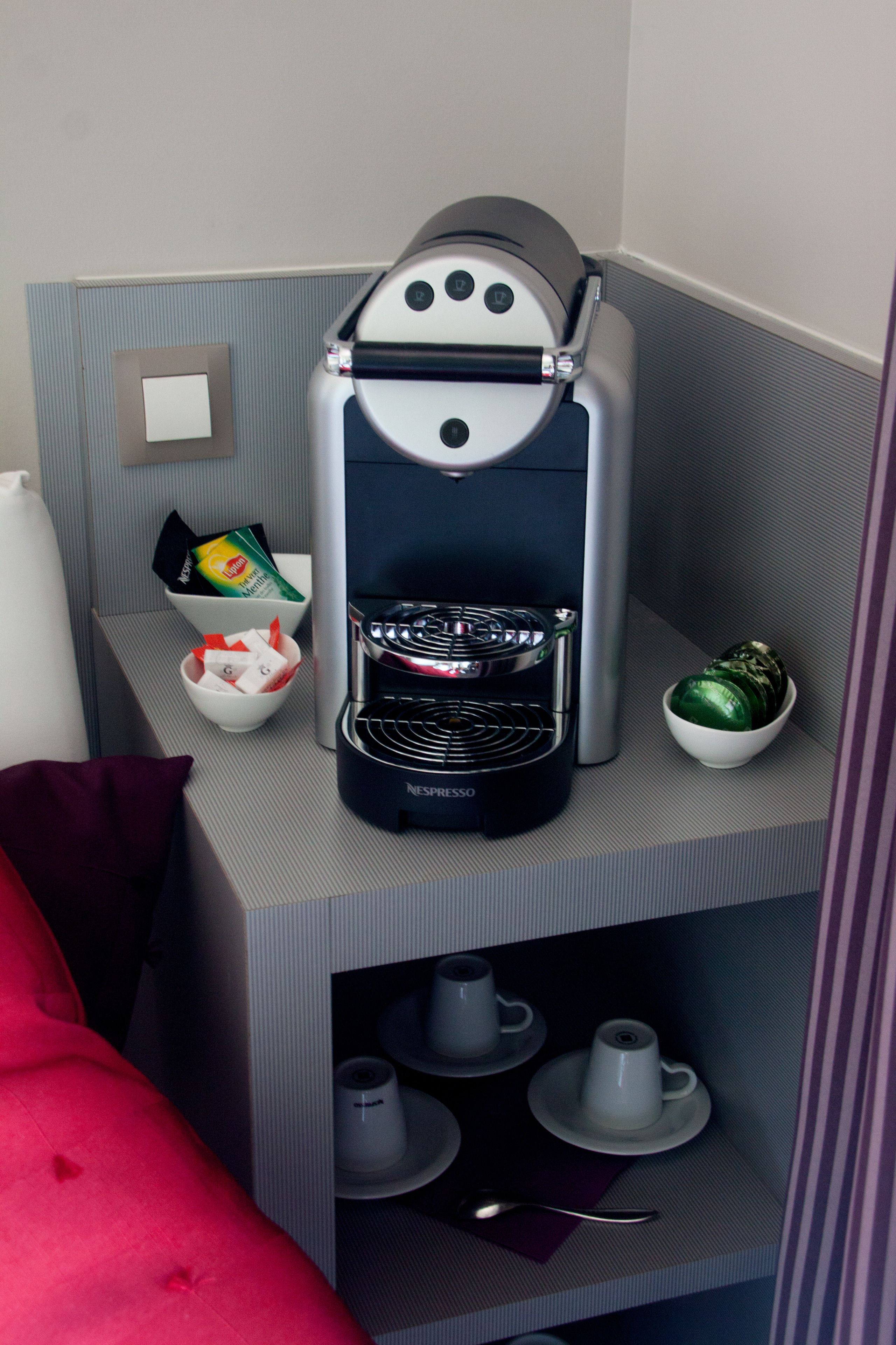 A Nespresso coffee machine on a gray countertop with a red pillow, a bowl of coffee pods, and a bowl of green coffee capsules.
