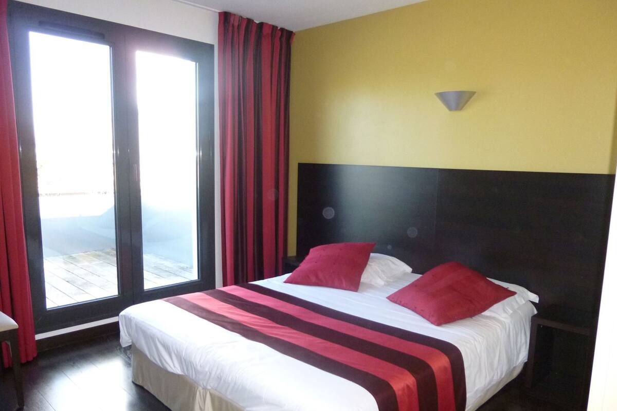 A hotel room with a bed, a window with curtains, a headboard, and a wall-mounted light fixture.
