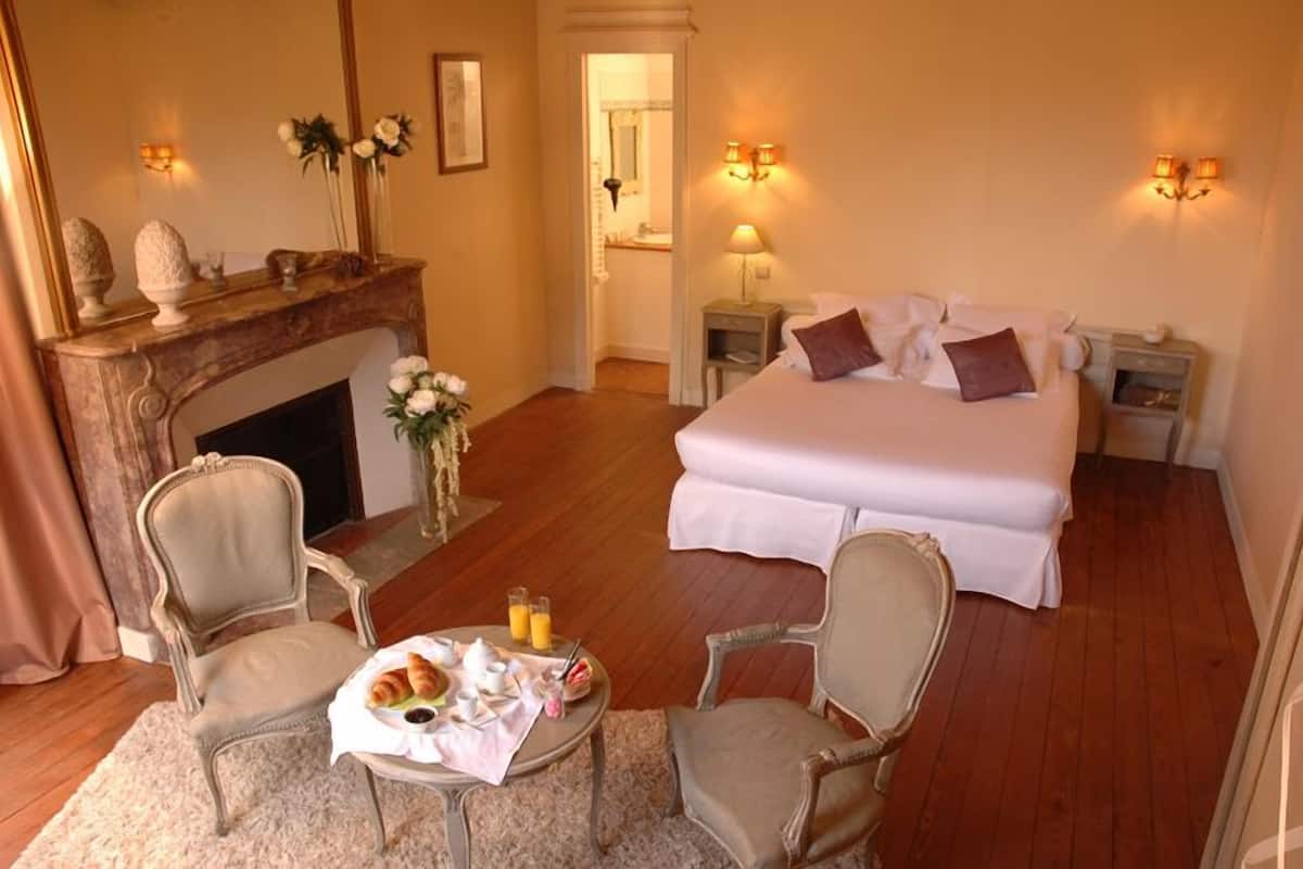 A hotel room with a large bed, two bedside tables, a fireplace, a sitting area with a table and chairs, and a mirror.