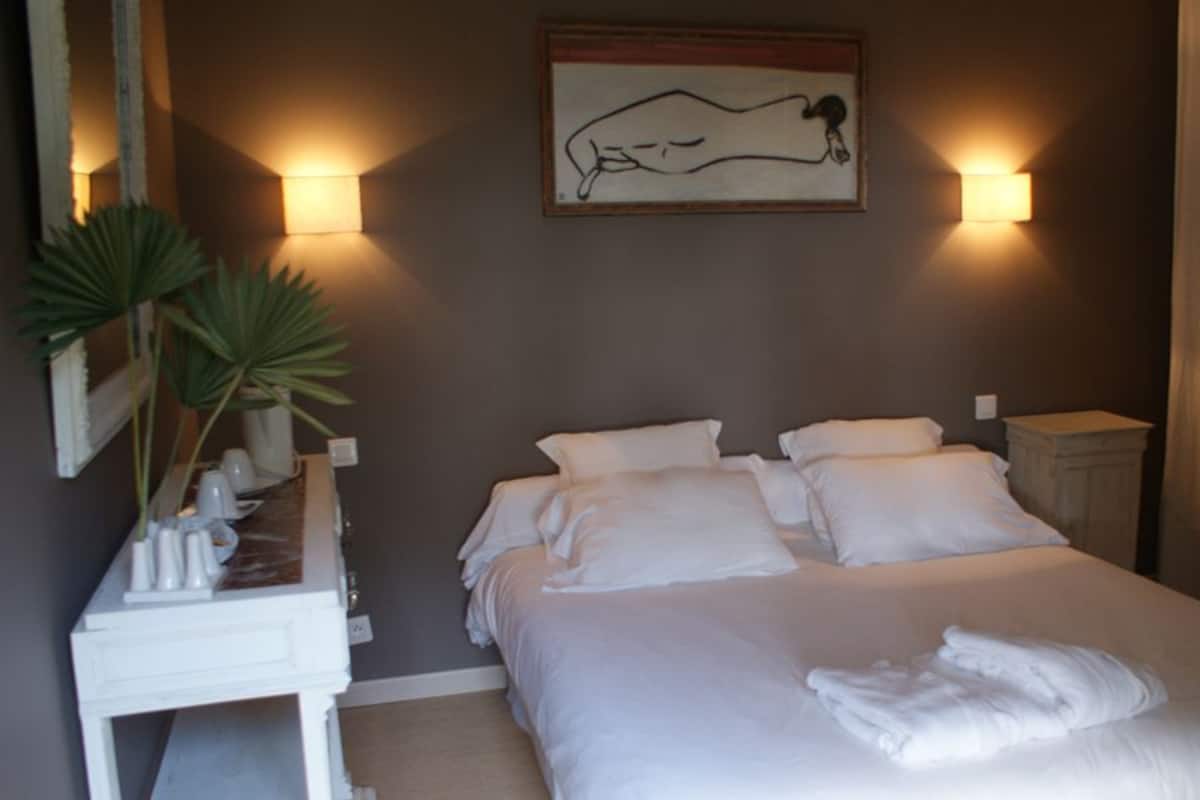 A bedroom with a large bed, two bedside tables, a nightstand, a painting on the wall, and a mirror.