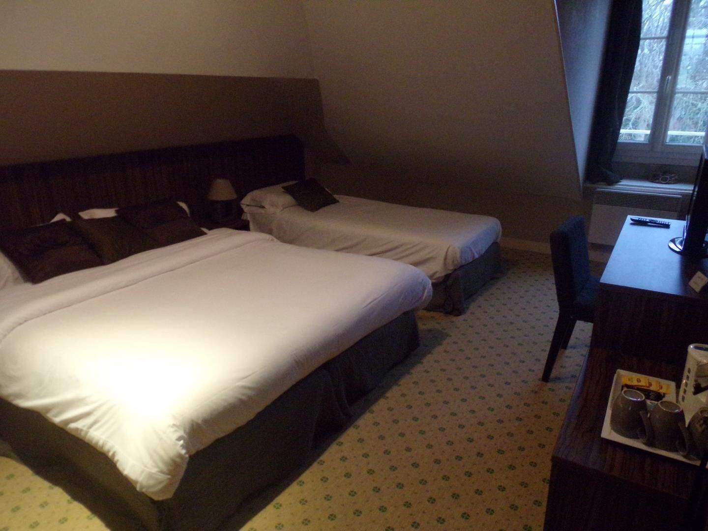 A hotel room with two single beds, a desk with a chair, and a window with a view of trees.