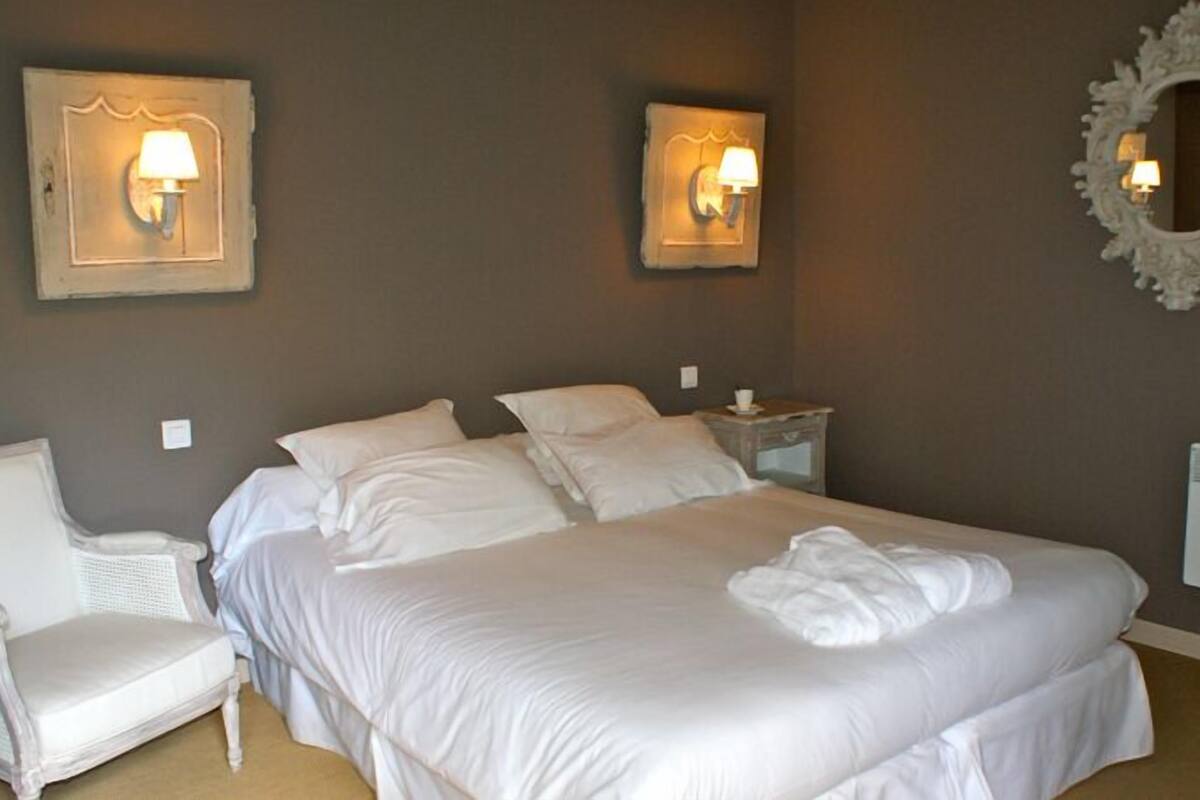 A neatly made bed with white linens, a white chair, and two wall-mounted lamps.