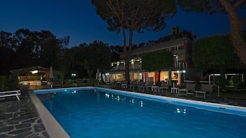 A swimming pool with lounge chairs and a building with multiple levels and outdoor lighting.