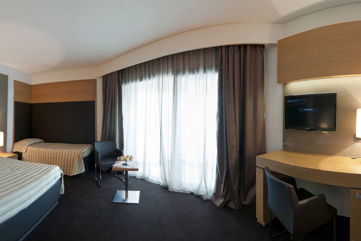 A hotel room with two beds, a desk, a chair, a TV, and a large window with curtains.