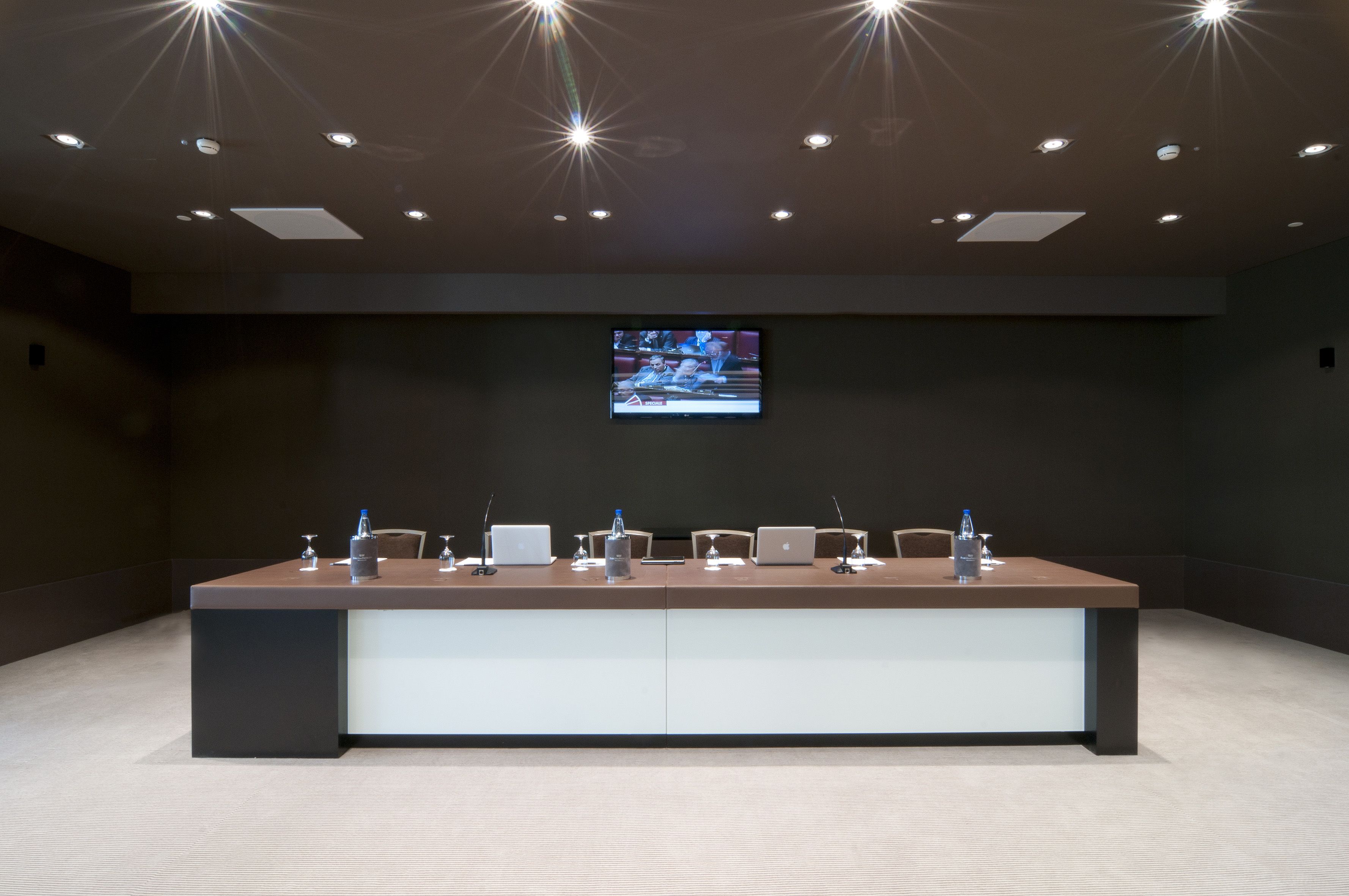A conference room with a long table, multiple chairs, a flat-screen TV, and recessed ceiling lights.