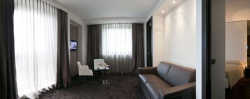 Una habitación de hotel moderna con alfombra oscura, un sofá gris, un sillón blanco, una mesita con un jarrón de flores y un televisor montado en la pared.