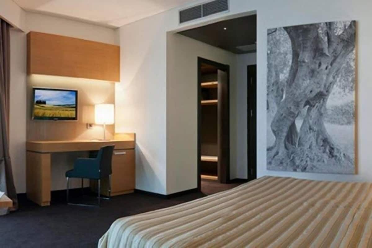 A hotel room with a bed, a desk, a chair, a television, and a painting on the wall.