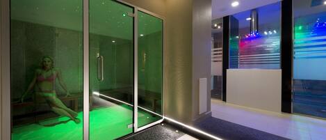 Une chambre d’hôtel moderne avec une cloison en verre, un espace de massage éclairé de lumière verte et une vue sur une installation lumineuse colorée à l’extérieur.