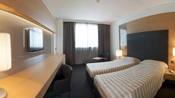 Ein Hotelzimmer mit zwei Betten, einem Schreibtisch, einem Sessel, einem Fernseher und einem Fenster mit Vorhängen.