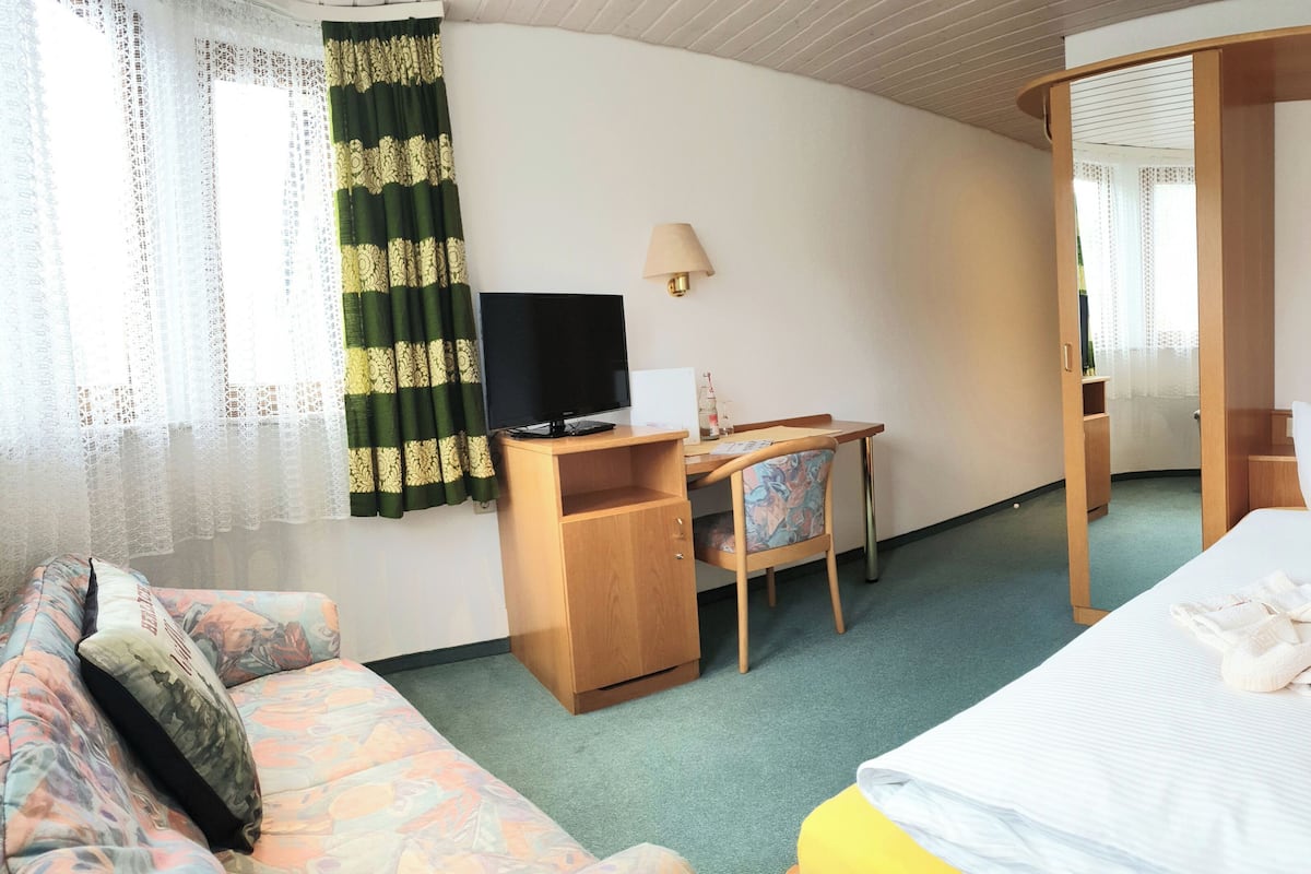 A hotel room with a bed, a desk with a chair, a television, and a window with curtains.