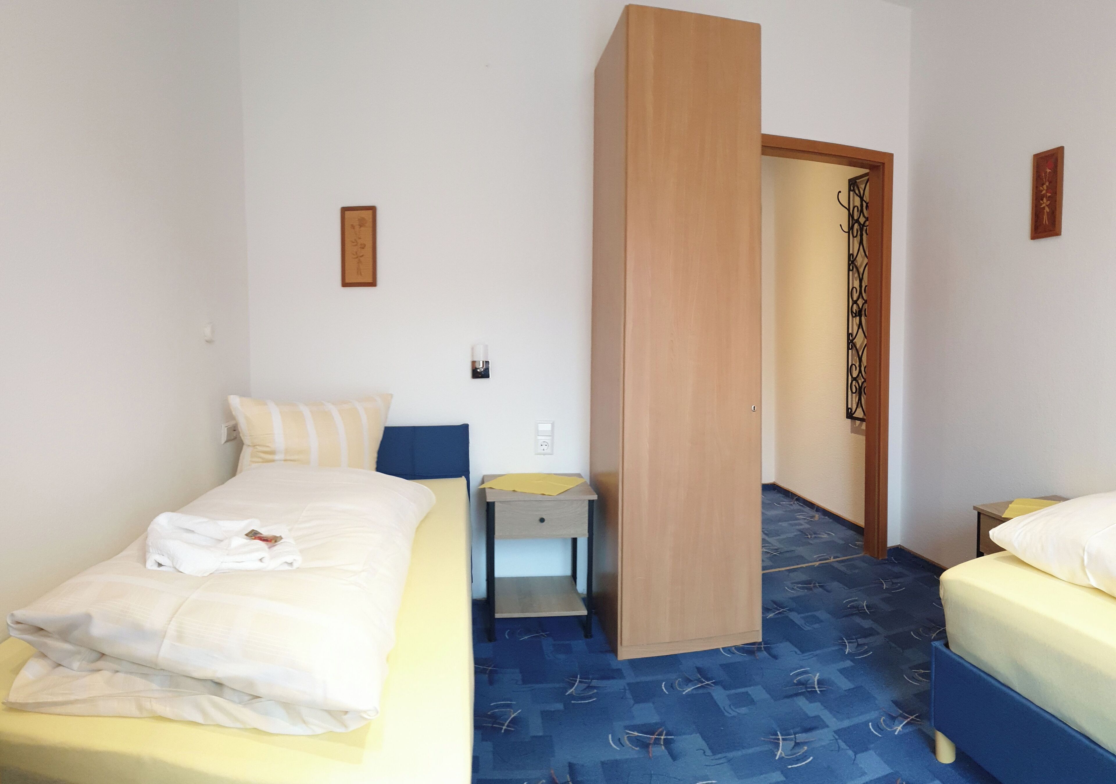 Ein Hotelzimmer mit zwei Betten, einem Holzschrank, einem kleinen Nachttisch und einem blau gemusterten Teppich.
