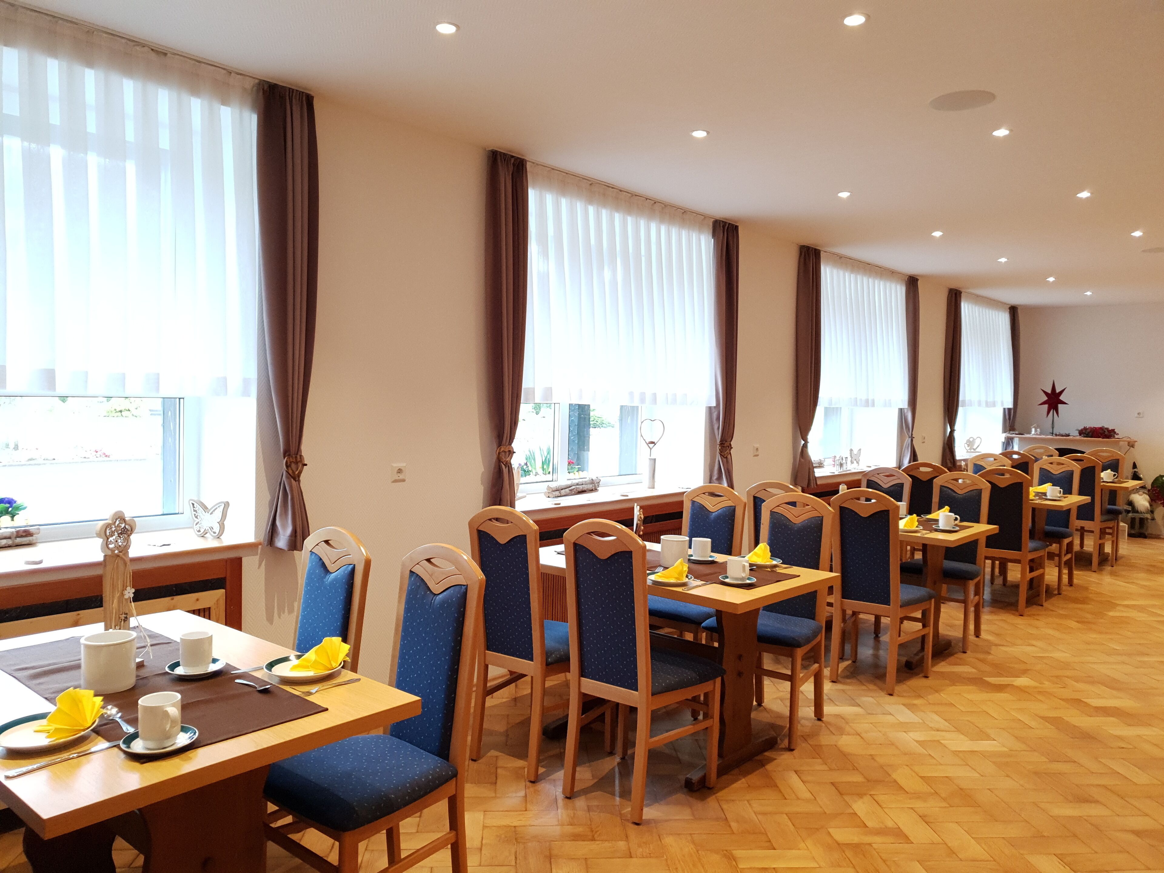 A dining area with multiple tables and chairs, large windows with curtains, and a wooden floor.