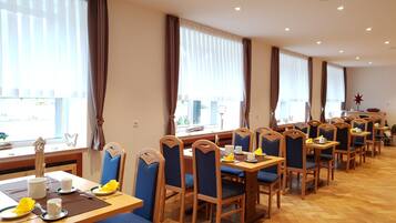 A dining area with multiple tables and chairs, large windows with curtains, and a wooden floor.