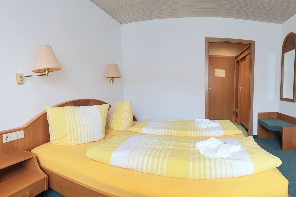 A hotel room with two beds, each with yellow bedding and striped pillows, a wooden headboard, a nightstand, a lamp, and a wooden door.