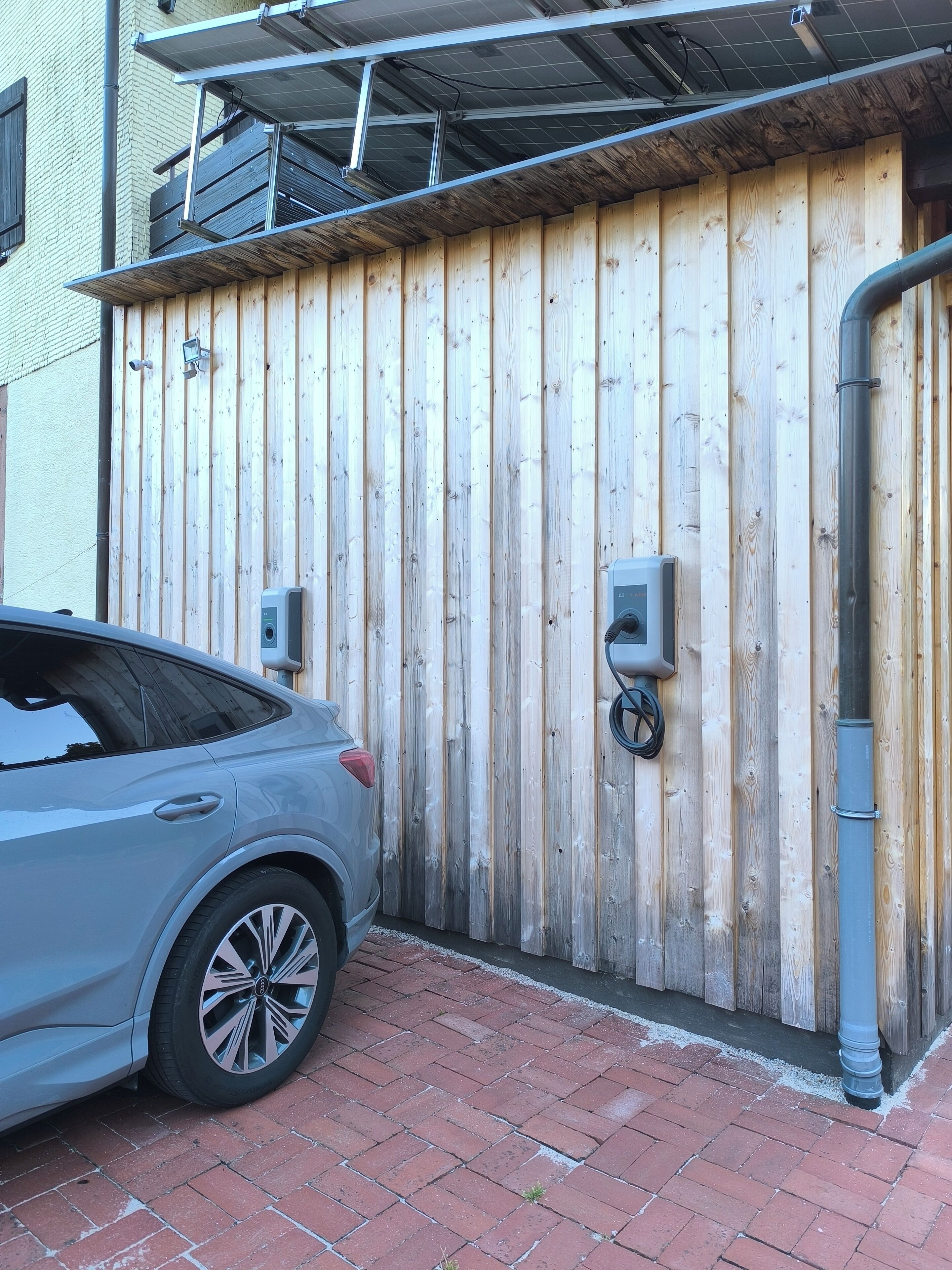 Un coche aparcado frente a una valla de madera con un portón metálico, un camino de ladrillo y un edificio con una ventana visible y un toldo de metal.