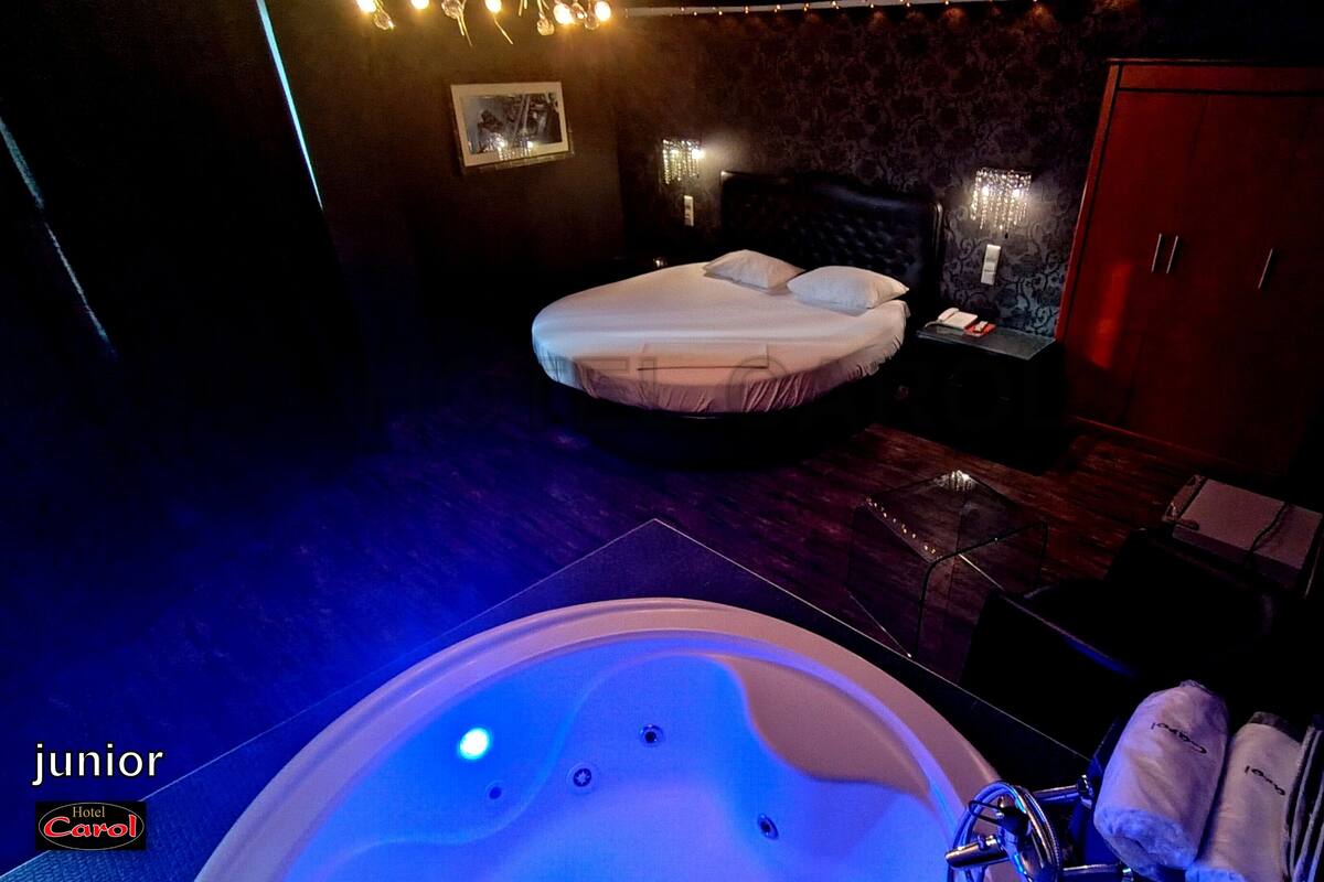 Un dormitorio tenuemente iluminado con una cama grande, suelo de madera, una mesita con una lámpara y un armario de madera.