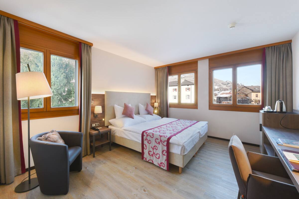 Habitación de hotel con cama, mesita de noche, lámpara, silla, escritorio y ventanas amplias que dan vistas a edificios y árboles.