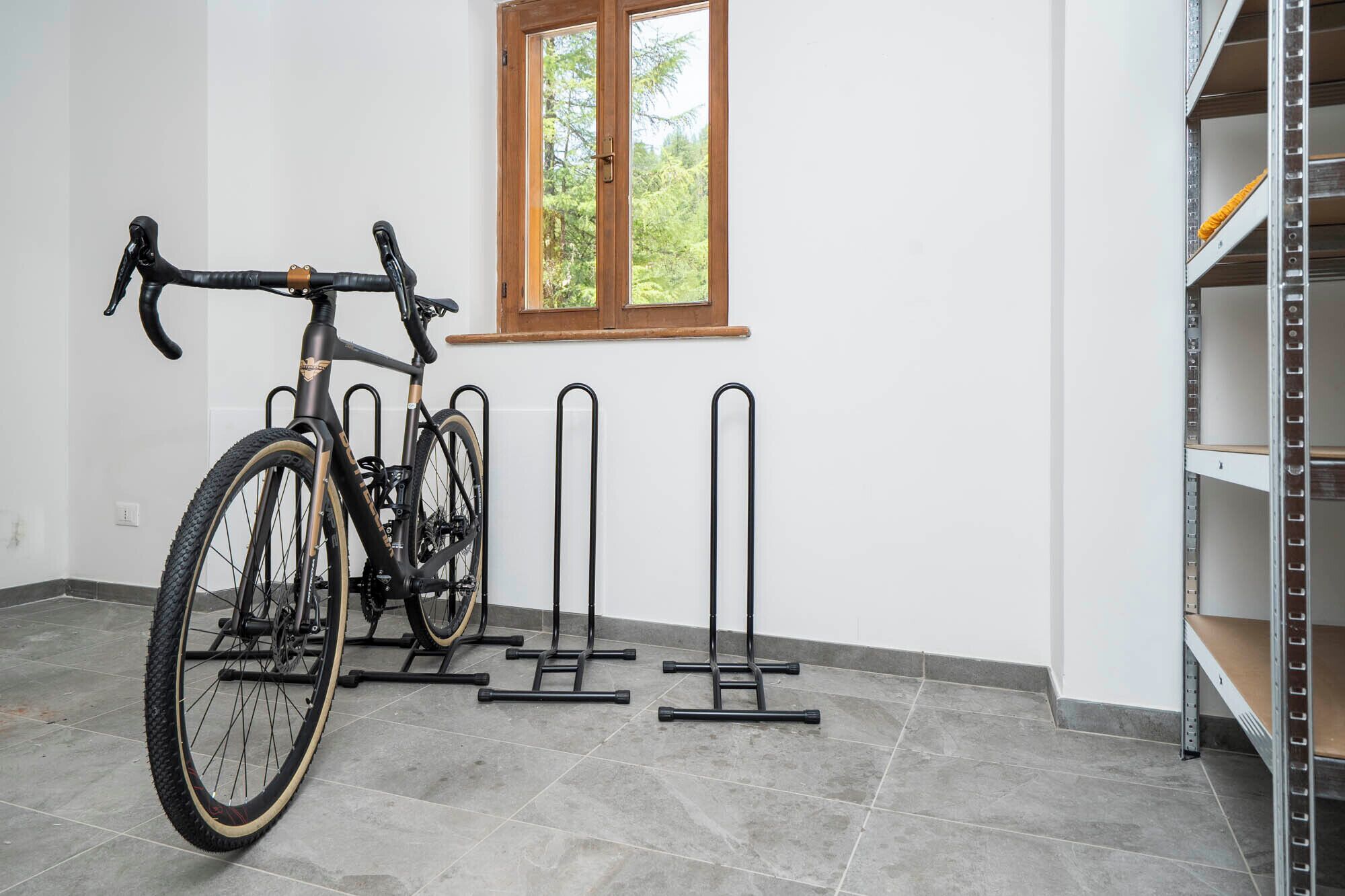 Una bicicleta sujeta a un soporte fijado a la pared en una habitación con suelo de baldosas, un ventanal con vistas a árboles y una estantería metálica.