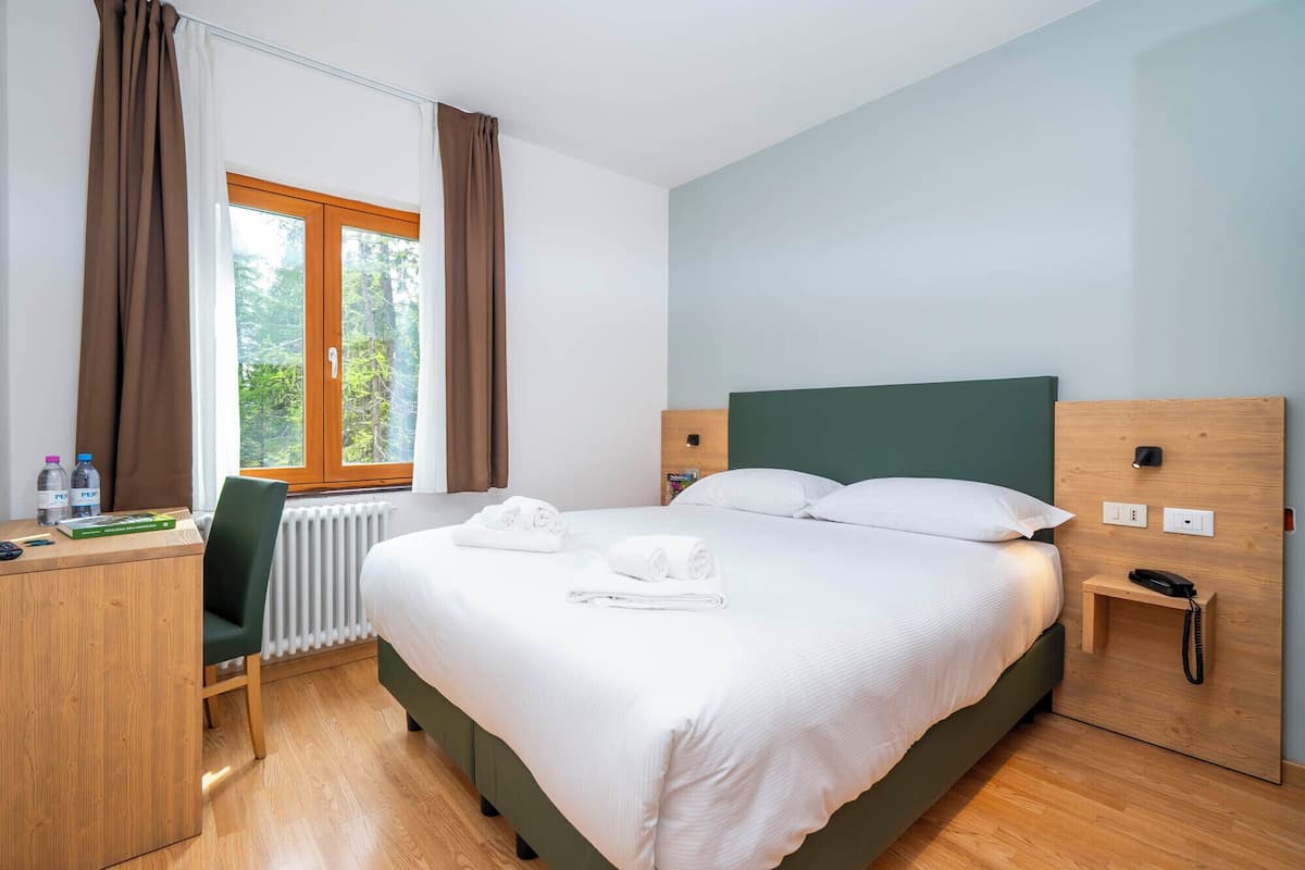 Une chambre d’hôtel comprenant un lit, un bureau avec une chaise, une table de chevet et une fenêtre avec des rideaux.