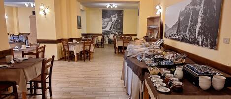 A dining area with wooden tables and chairs, a buffet with various food items, and a large mountain landscape painting on the wall.