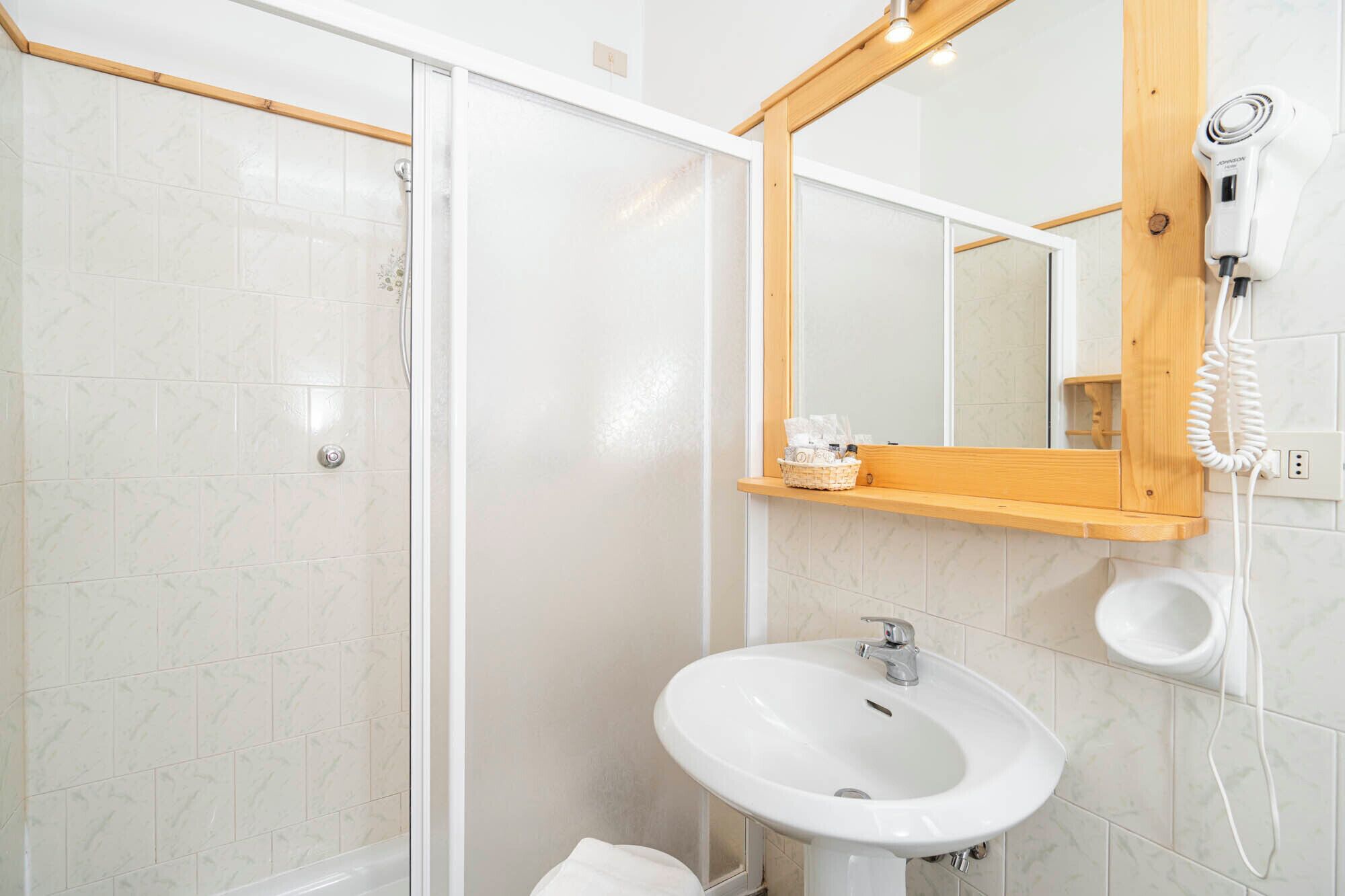 A bathroom with a glass shower door, a white sink, a wooden shelf, and a hairdryer mounted on the wall.
