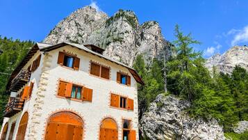 A stone building with wooden shutters and balconies, set against a mountainous backdrop.